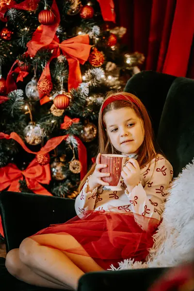 Süslü bir Noel ağacının yanındaki sandalyede oturan genç bir kız elinde kırmızı çizgili bir kupa, sıcacık ve neşeli bir bayram anının tadını çıkarıyor..