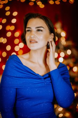 Pırıl pırıl mavi elbiseli zarif kadın, arka planda altın bokeh ışıkları ve kırmızı dekoru olan bir şenliğe karşı güvenle poz veriyor..