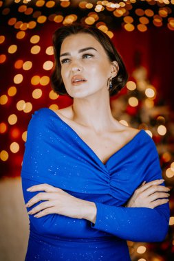 Elegant woman in a sparkling blue dress poses confidently against a festive background with golden bokeh lights and red decor. clipart