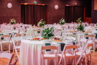 Yemek masaları yuvarlak, beyaz masa örtüleri, çiçek süslemeleri ve ahşap döşeme üzerine düzenlenmiş sofra takımı olan zarif etkinlik salonu..