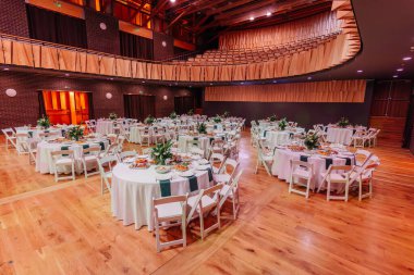 Beyaz masa örtüleri, yeşil peçeteler ve ahşap zemin üzerindeki çiçek süslemeleriyle süslenmiş yuvarlak masalı ziyafet salonu..