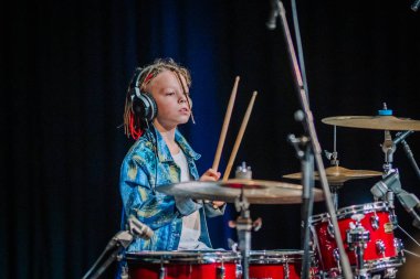 Cesis, Latvia - December 5, 2024 - A young drummer with headphones playing a red drum set on stage during the Rairu 25 event, showcasing musical talent and concentration. clipart