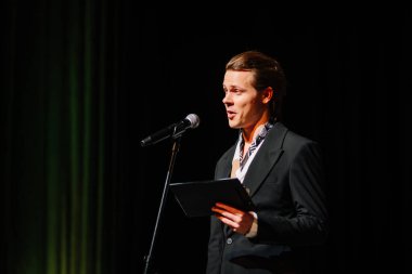 Cesis, Letonya - 5 Aralık 2024 - Sahnede takım elbiseli bir adam mikrofona konuşuyor, elinde tablet, karanlık bir arka plan ve odaklanmış sahne aydınlatması var. Rairu 25