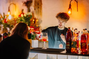 Valmiera, Latvia - December 7, 2024 - A bartender pours a vibrant cocktail at a warmly lit bar, surrounded by bottles and floral decor. Warm ambiance and customer interaction. clipart