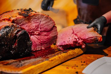 Valmiera, Letonya - 7 Aralık 2024 - Ahşap bir kesme tahtasının üzerinde taze dilimlenmiş, sulu bir et, şeflerin eldivenli elleri ve oyma bıçağı görünürde