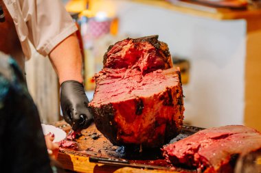 Valmiera, Letonya - 7 Aralık 2024 - Ahşap bir kesme tahtasının üzerinde taze dilimlenmiş, sulu bir et, şeflerin eldivenli elleri ve oyma bıçağı görünürde