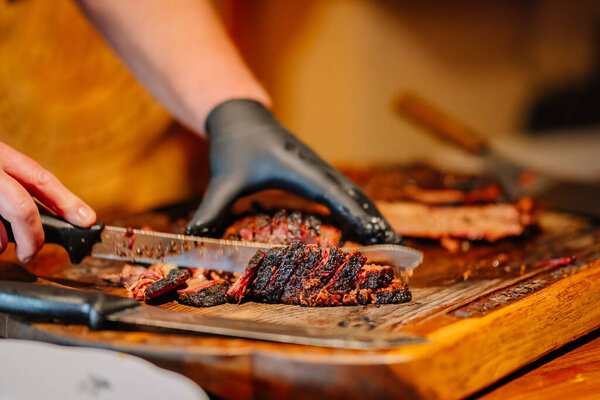 Valmiera, Latvia - December 7, 2024 - A close-up of a person slicing smoked, charred meat with a knife on a wooden cutting board, wearing black gloves for handling food.