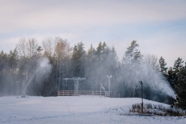 Snowmaking machines in operation at a ski slope surrounded by a winter forest under a cloudy blue sky. clipart