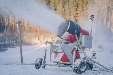 Kırmızı bir kar topu kayak yamacına yapay kar püskürtüyor. Etrafı karlı araziler ve arka plandaki ağaçlarla çevrili..