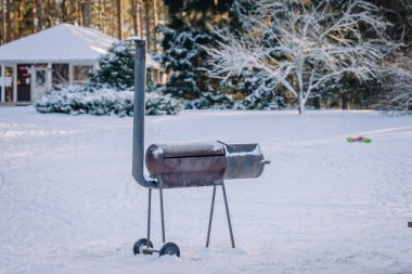Valmiera, Letonya - 4 Ocak 2025 - Karlı bir parkta karla kaplı bir ızgara, ağaçlarla çevrili ve arka planda bir ev, soğuk bir kış günü.