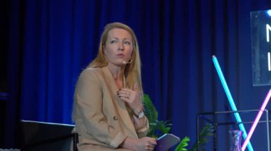 Valmiera, Letonya - 18 Ekim 2024 - Bir yapay zeka konferansında kadın bir konuşmacı, elinde bir tablet, mikrofonla konuşuyor, arka planda neon ışıkları ve bitkilerle sahnede oturuyor.