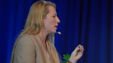 Valmiera, Letonya - 18 Ekim 2024 - Yapay Zeka konferansında bej ceket giyen, kulaklıkla konuşan, mavi arka planı ve parlak ışıkları olan bir kadın konuşmacı.