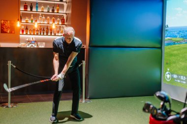 Riga, Letonya - 8 Ocak 2024 - Bir golfçü arka planda bir çubuğu ve aydınlatılmış rafları olan kapalı bir golf simülatöründe sallanmaya hazırlanıyor.