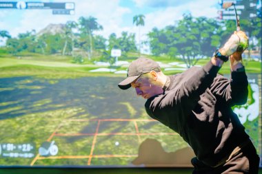 Riga, Letonya - 8 Ocak 2024 - Siyah kıyafetli odaklanmış bir golfçü, arka planda yemyeşil manzaralı sanal bir simülatör ekranının önünde golf sopasını sallıyor.