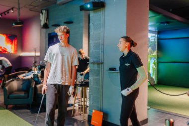 Riga, Letonya - 8 Ocak 2024 - Golf eldivenli genç bir adam ve bir kadın kapalı bir golf salonunda sohbet veya antrenman oturumunda bulunuyorlar..