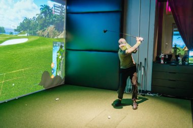 Riga, Letonya - 8 Ocak 2024 - Bir golfçü ekranda manzaralı sanal bir kursla kapalı bir golf simülatöründe güçlü bir vuruş yapıyor.