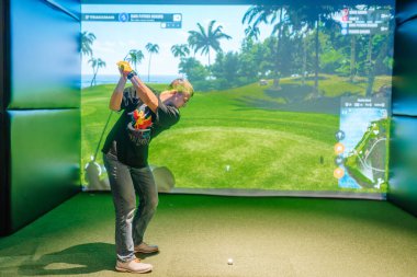 Riga, Letonya - 8 Ocak 2024 - Kapalı bir Trackman simülatöründe sallanan bir golfçü, ekran üzerinde palmiye ağaçları ve tropikal bir çevre bulunan yemyeşil bir sanal golf sahası.