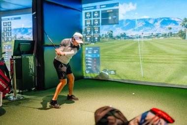 Riga, Letonya - 8 Ocak 2024 - İçerde Trackman simülatörü kullanan bir golfçü, ekranda sanal golf sahası ve performans verileri olan bir sopayı sallıyor.