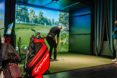 Riga, Letonya - 8 Ocak 2024 - TaylorMade golf çantası ve kapalı bir pist simülatöründe golf sopaları, ekranda sanal bir golf sahası sergilenmeye hazırlanan bir oyuncu.