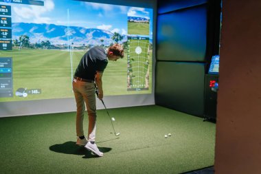 Riga, Letonya - 8 Ocak 2024 - İçerde Trackman simülatörü kullanan bir golfçü, topa sanal bir golf sahası ve ekrandaki performans verileriyle vurmaya odaklanıyor.