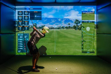 Riga, Letonya - 8 Ocak 2024 - İçerde Trackman simülatörü kullanan bir golfçü, ekranda sanal golf sahası ve performans verileri olan bir sopayı sallıyor.