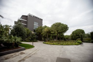 Belek, Türkiye - 22 Şubat 2020 - Çağdaş bina yemyeşil ağaçlar, peyzaj bahçesi ve bulutlu bir gökyüzü altında beton bir yol ile çevrili.