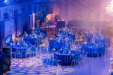 Riga, Letonya - 11 Ocak 2025 - Yuvarlak masalar, mavi dekorasyonlar, şeffaf sandalyeler ve yumuşak ışıklandırma ile lüks ve şenlikli bir ortam yaratan zarif bir etkinlik salonu.