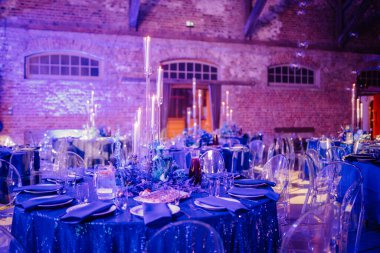 Mavi pullu masa örtüsü, uzun mumlar, çiçekli süs, cam sandalyeler ve tuğladan duvarlı bir mekanda yumuşak ortam aydınlatması olan zarif bir etkinlik masası..