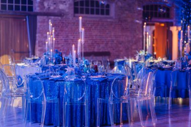 Uzun mumlar, mavi çiçekli süsler ve güzel ışıklandırılmış bir salonda şeffaf sandalyeler..