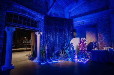 Mercan şekilli sualtı temalı dekoratif düzenek, mavi pullu arka plan, beyaz sütunlar, ve bir etkinlik salonundaki yumuşak parlak ışıklar.