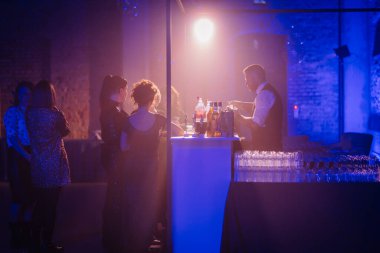 Riga, Letonya - 11 Ocak 2025 - Bir grup kadına, atmosferik mavi ve ılık ışıklandırmalı loş bir barda içki servisi yapan bir barmen.