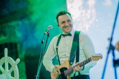 Riga, Letonya - 11 Ocak 2025 - Buz Diyarı temalı bir etkinlik sırasında beyaz gömlekli ve pantolon askısı takmış gülümseyen bir gitarist, elinde elektro gitaristle sahneye çıktı..