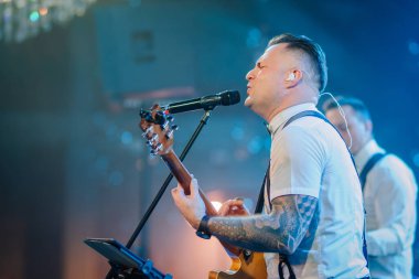 Riga, Letonya - 11 Ocak 2025 - Kolu dövmeli bir müzisyen Ice Land temalı bir etkinlikte gitarıyla sahnede sahne ışıkları altında bir mikrofona şarkı söylüyor..