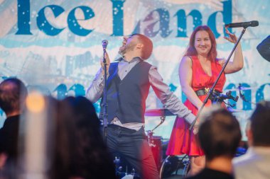 Riga, Letonya - 11 Ocak 2025 - Buz Diyarı temalı bir etkinlikte kırmızı elbiseli neşeli bir kadınla coşkulu bir şekilde sahnede performans sergileyen bir adam.