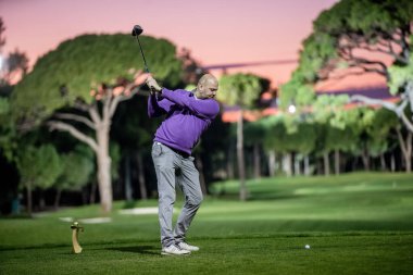 Türkiye, Belek - 24 Şubat 2020 - Mor kazaklı bir golfçü, günbatımında yeşil bir pistte, arka planda ağaçlar ve sakin bir gökyüzü olan bir golf sopasını sallıyor.