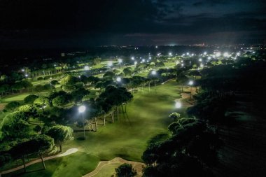 Belek, Türkiye - 24 Şubat 2020 - Parlak spot ışıkları, yemyeşil otlaklar, ağaçlar ve uzakta sakin bir şehir manzarasının yer aldığı aydınlık bir golf sahasının havadan görünüşü.