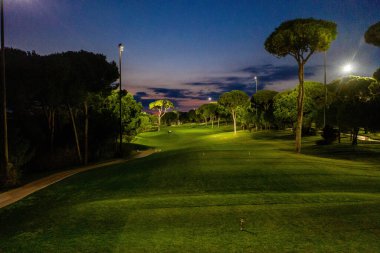 Belek, Turkey - February 24, 2020 - A brightly lit golf course at twilight with lush green fairways, tall trees, and dramatic evening skies creating a tranquil atmosphere. clipart