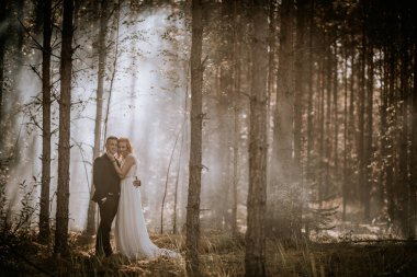 Valmiera, Letonya - 24 Ağustos 2024 - Bir gelin ve damat, ağaçların arasından akan güneş ışığıyla çevrili sisli bir ormanda, romantik ve huzurlu bir atmosfer yaratarak dururlar..