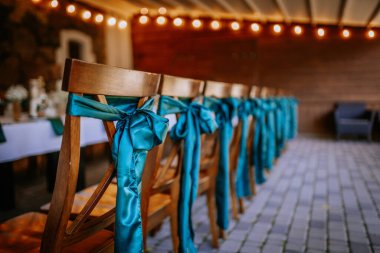 Arkasında mavi saten kurdeleler olan ahşap sandalyeler, dekore edilmiş bir etkinlik salonunda, sıcak ışıklandırma ve bulanık arka planda..