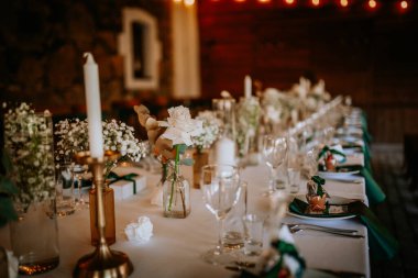 Beyaz masa örtüsü üzerinde çiçekler, mumlar ve hediye kutuları olan zarif bir düğün masası romantik ve sofistike bir atmosfer yaratıyor..