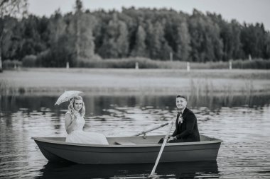 Dantel şemsiyeli bir gelin ve sakin bir gölde kayıkta bir damat, sakin doğal bir arka planla siyah beyaz yakalanmış..