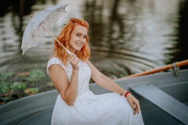 Valmiera, Letonya - 24 Ağustos 2024 - Beyaz bir gelinlik içinde dantel bir şemsiye tutan, sakin bir gölde kayıkta oturan, arka planda su dalgalanmaları olan bir gelin.