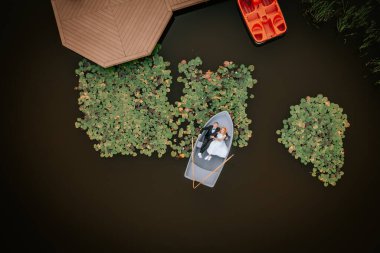 Aerial view of a couple in wedding attire lying in a rowboat on a pond with lily pads and a dock, surrounded by tranquil water. clipart