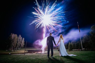 Gelin ve damat el ele tutuşuyor, göz kamaştırıcı bir havai fişek gösterisine bakıyor açık havada romantik bir gökyüzü altında.