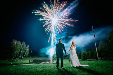 Gelin ve damat el ele tutuşuyor, göz kamaştırıcı bir havai fişek gösterisine bakıyor açık havada romantik bir gökyüzü altında.