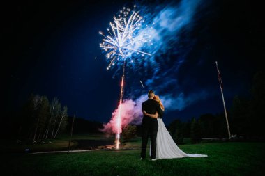 Gelin ve damat, yıldızlı bir gökyüzünde canlı bir havai fişek gösterisinin altında, ağaçlarla çevrili ve romantik bir ortamda bayrak direğiyle sarılıyorlar..