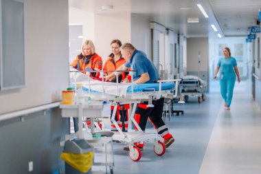 Valmiera, Letonya - 17 Ocak 2025 - Turuncu üniformalı acil tıp personeli hastane koridorunda bir hastaya sedyede yardım ediyor. Arka planda başka bir hemşire yürüyor..