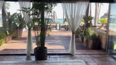 Sotogrande, İspanya - 24 Ocak 2024 - Güzel bir sahil bölgesinde huzurlu bir ortam sunan akarsu perdeleri, tropikal bitkiler ve okyanus manzaralı sakin bir teras.