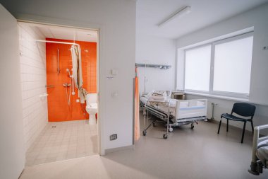 Valmiera, Letonya - 17 Ocak 2025 - Yataklı bir hastane odası, bebek beşiği, sandalye, büyük pencereler ve hasta bakımı için tasarlanmış portakal renkli banyolar.