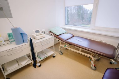 Valmiera, Letonya - 17 Ocak 2025 - Kahverengi tıbbi yataklı, teşhis ekipmanlı ve pencereye bakan temiz ve iyi ışıklandırılmış klinik ortamlı muayene odası.
